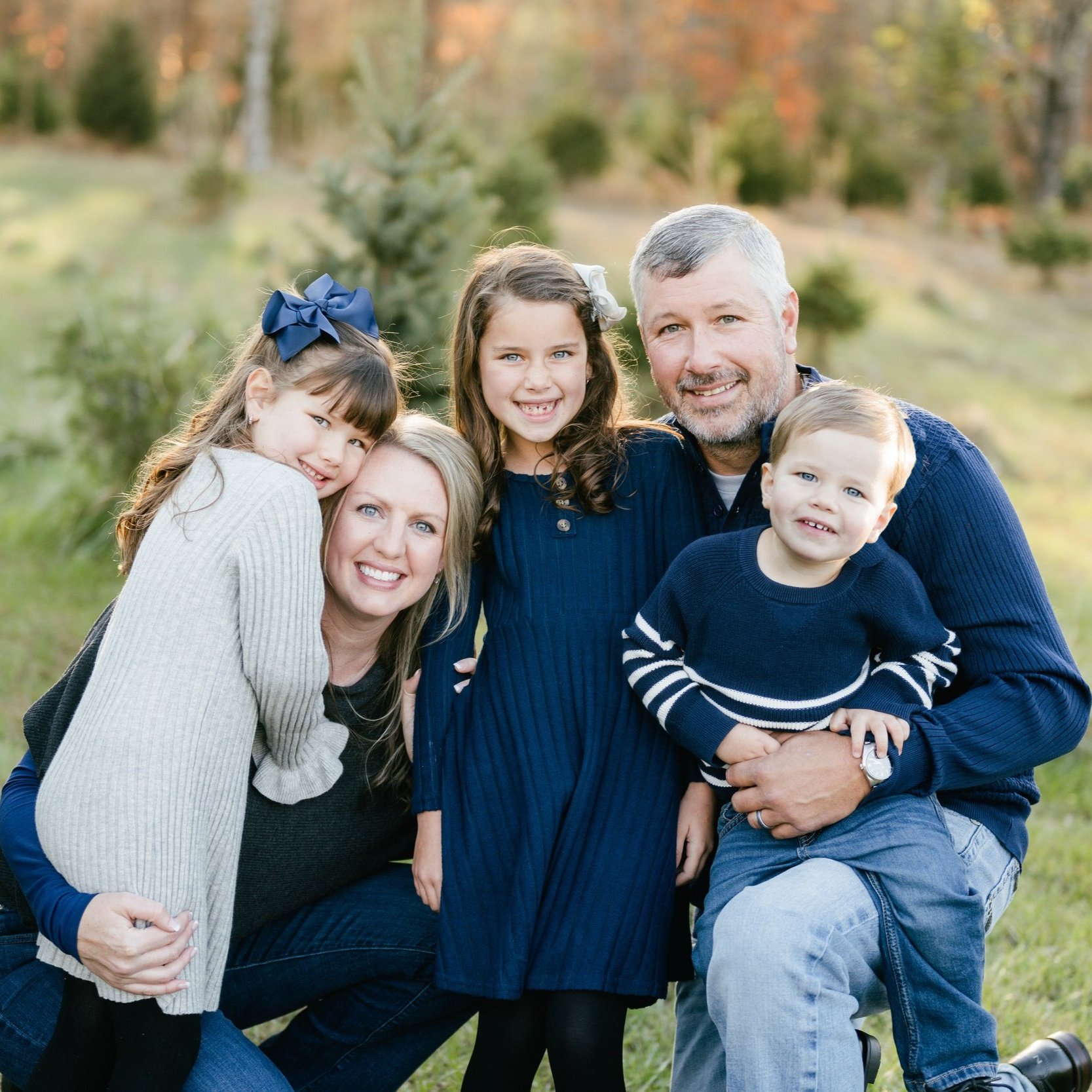 Family portrait of a smiling group in a natural outdoor setting, featuring a mother, father, and three children dressed in coordinated outfits. The scene captures a joyful moment among lush greenery, highlighting family togetherness and warmth.